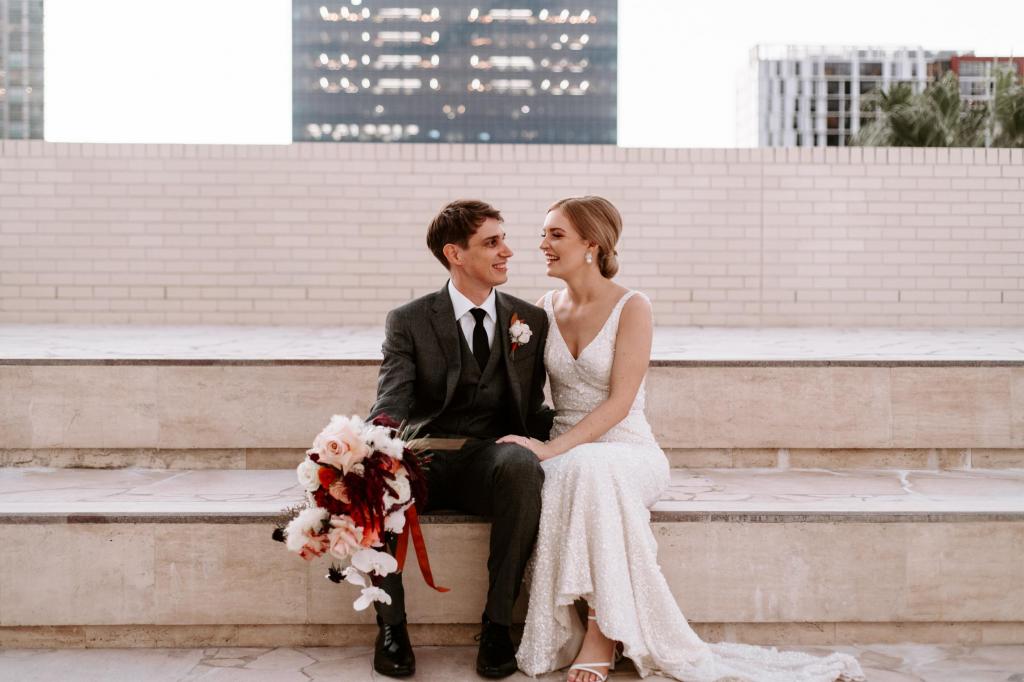 KWH real bride Katie sits with her husband on cement steps in her simple yet glamorous Lola wedding dress with train.