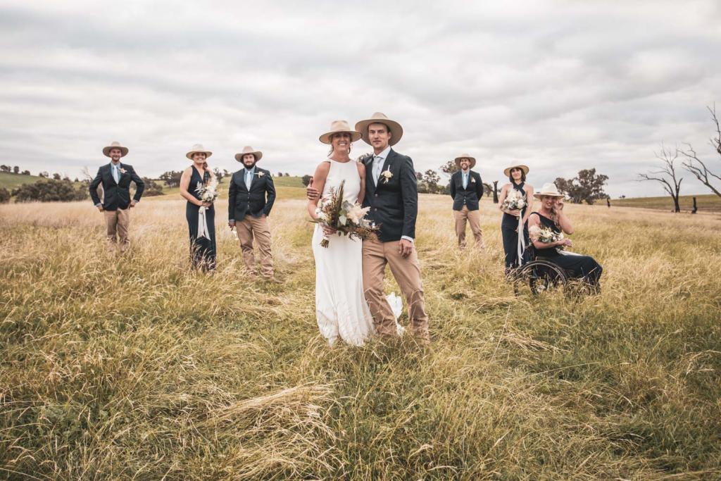 KWH real bride Angela stands with Graham in front of their wedding party in a field. She wears the simple Bridget gown with waist cutouts.