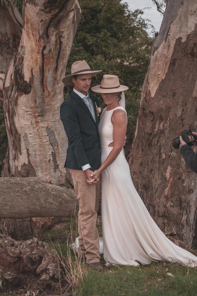 KWH real bride Angela and Graham holding hands by a large tree. She wears the fitted Bridget gown with waist cutouts.
