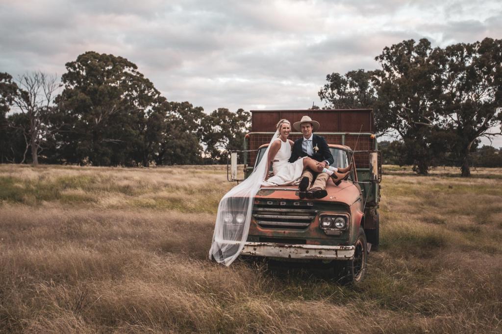 KWH real bride Angela and Graham sit on top of an old Dodge truck in the field. She wears the bold Bridget gown with high neckline.