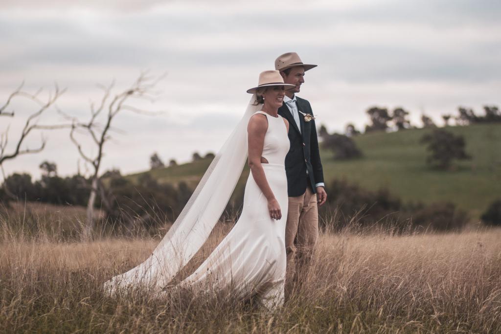 KWH real bride Angela walks in the field with her husband Graham. They both wear Akubra hats and she wears the simple Bridget gown.