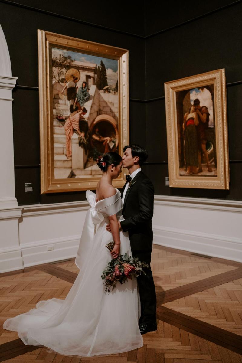 KWH real bride Sarah standing with Brian looking at a red gallery wall. She wears the traditonal Kitty Joni gown with a-line skirt and fitted bodice.