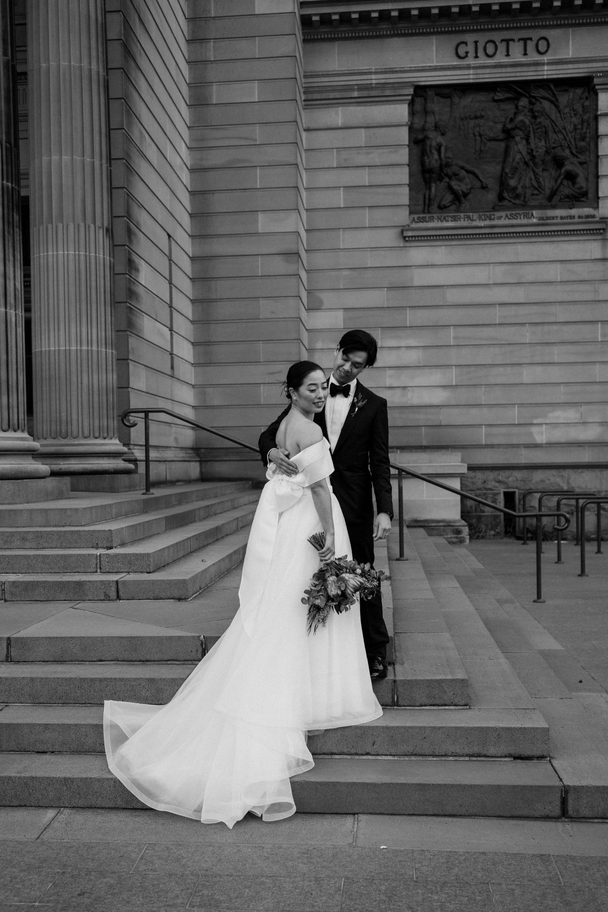 B&W photo of Sarah and brian out front of the Art Gallery. She wears the custom Kitty Joni gown with long train and bow in the back.