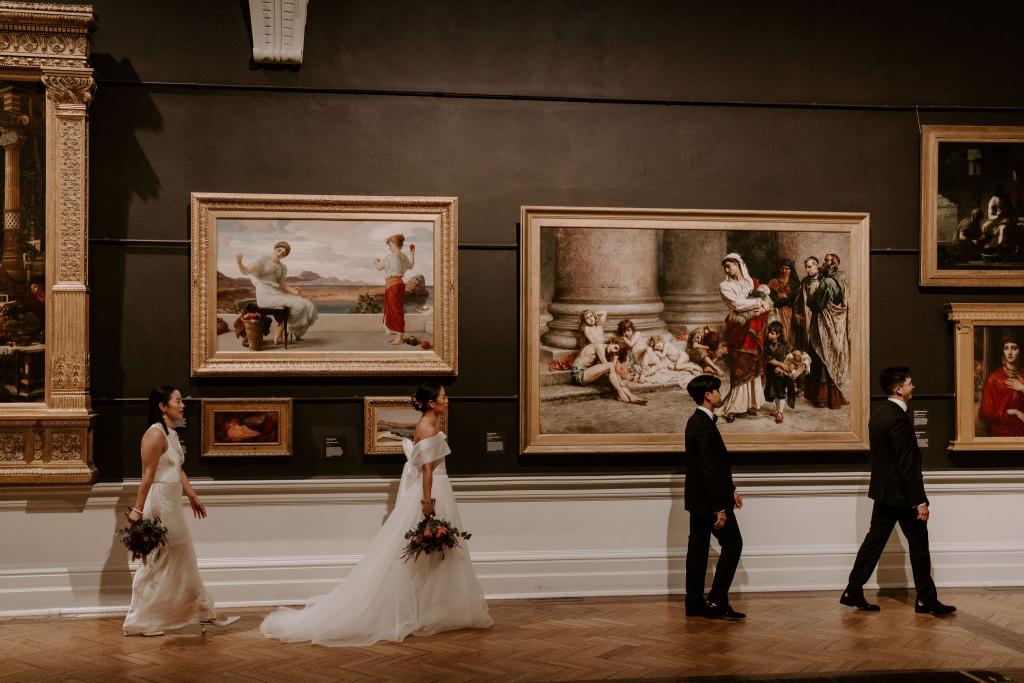 KWH real bride Sarah and Brian with their bridal party walk past large paintings in the gallery. She wears the ivory strapless Kitty Joni gown.