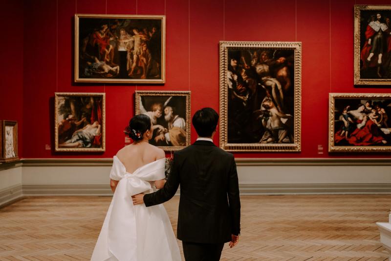 KWH real bride Sarah standing with Brian looking at a red gallery wall. She wears the traditonal Kitty Joni gown with a-line skirt and fitted bodice.
