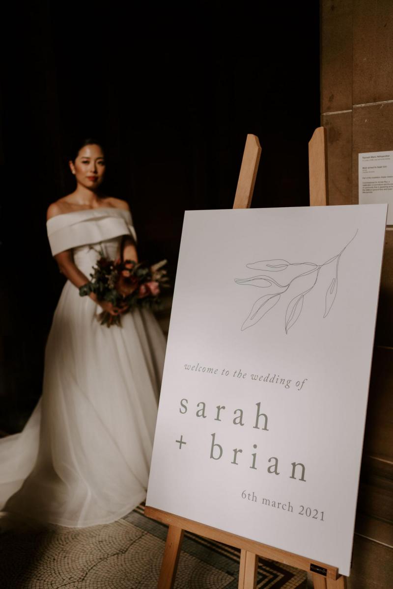 KWH real bride Sarah stands behind the welcome sign to the ceremony. She wears the bespoke Kitty Joni gown.