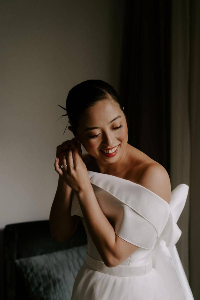 KWH real bride Sarah attaching her earring while wearing the Kitty Joni gown with twill and tulle fabrics.