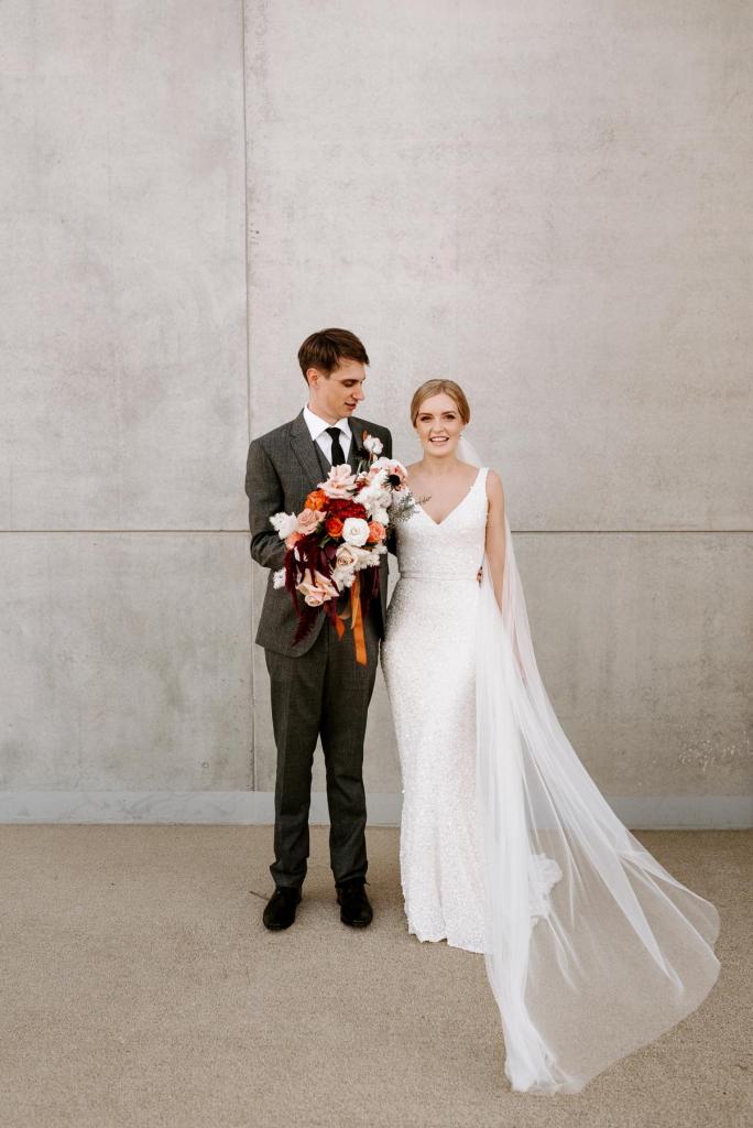 KWH real bride Katie and Adrian stand against a cement wall. She wears the minimalist Lola gown with fit and flare shape.