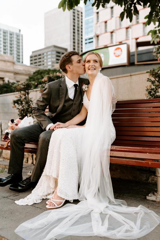 KWH real bride Katie sits with her husband on a park bench as he kisses her cheek. She wears the stunning ivory Lola wedding dress with sequins and matching belt.