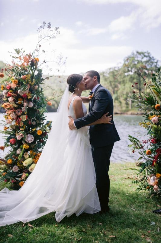 KWH real bride Annalese kisses her new husband at their colorful floral alter. She wears the modern Aisha gown with aline skirt and mesh fabric.