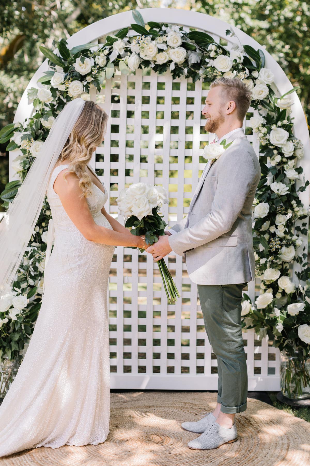 KWH real bride Bec and Nick stand at a white lattic alter. She wears the beaded Olympia wedding dress with organic pattern.