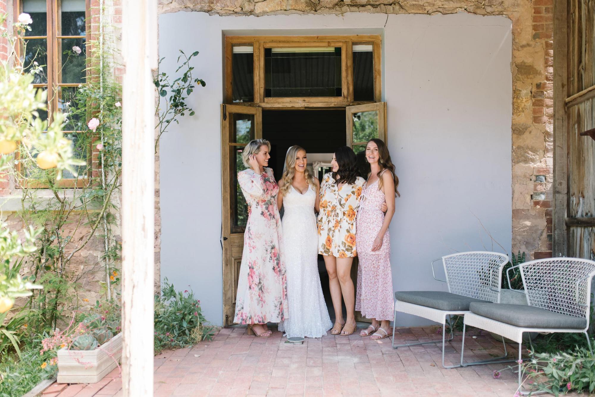 KWH real bride stands with her bridesmaids in front of a door. She wears the fit and flare Olympia beaded wedding dress.