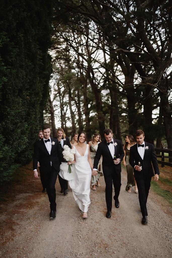 Real bride Charlotte with the groomsmen walking down a tree lined street. She wears the simple Leonie Alexia gown by KWH