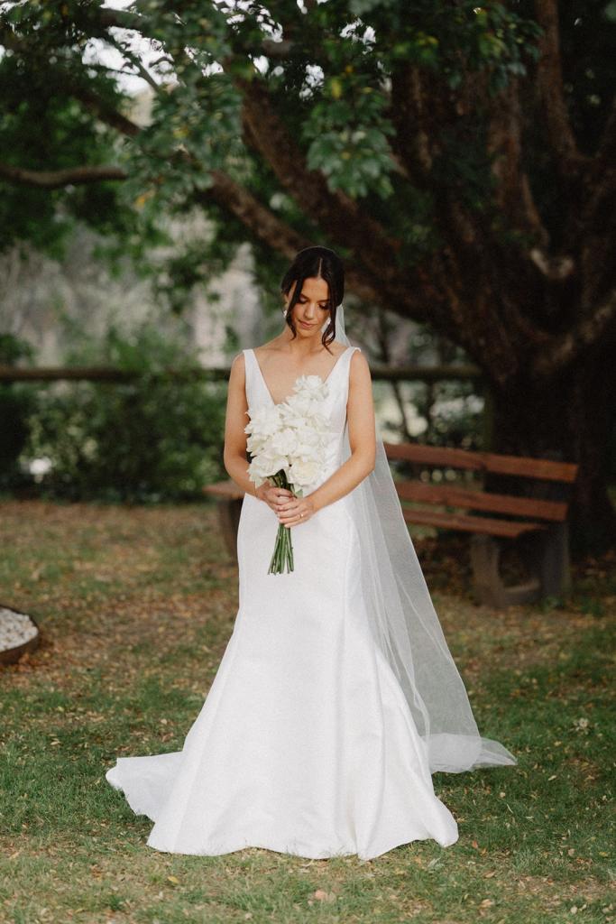 Real bride Charlotte stands with her white bridal bouquet by Sammy Bate. She wears the timeless Leonie Alexia gown with U-neck.