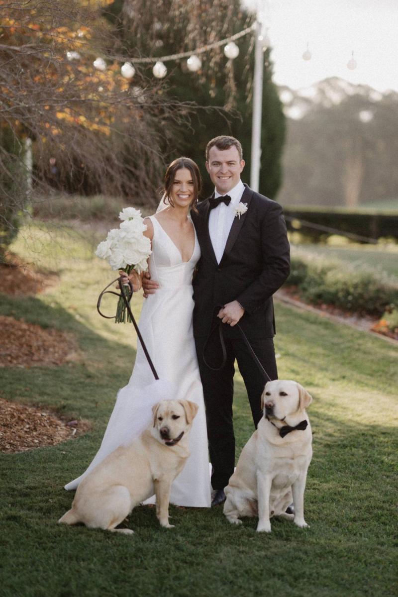 Real bride Charlotte and Myles taking photos with their two blond labs who are wearing black bowties. She wears the elegant Leonie Alexia gown by KWH.