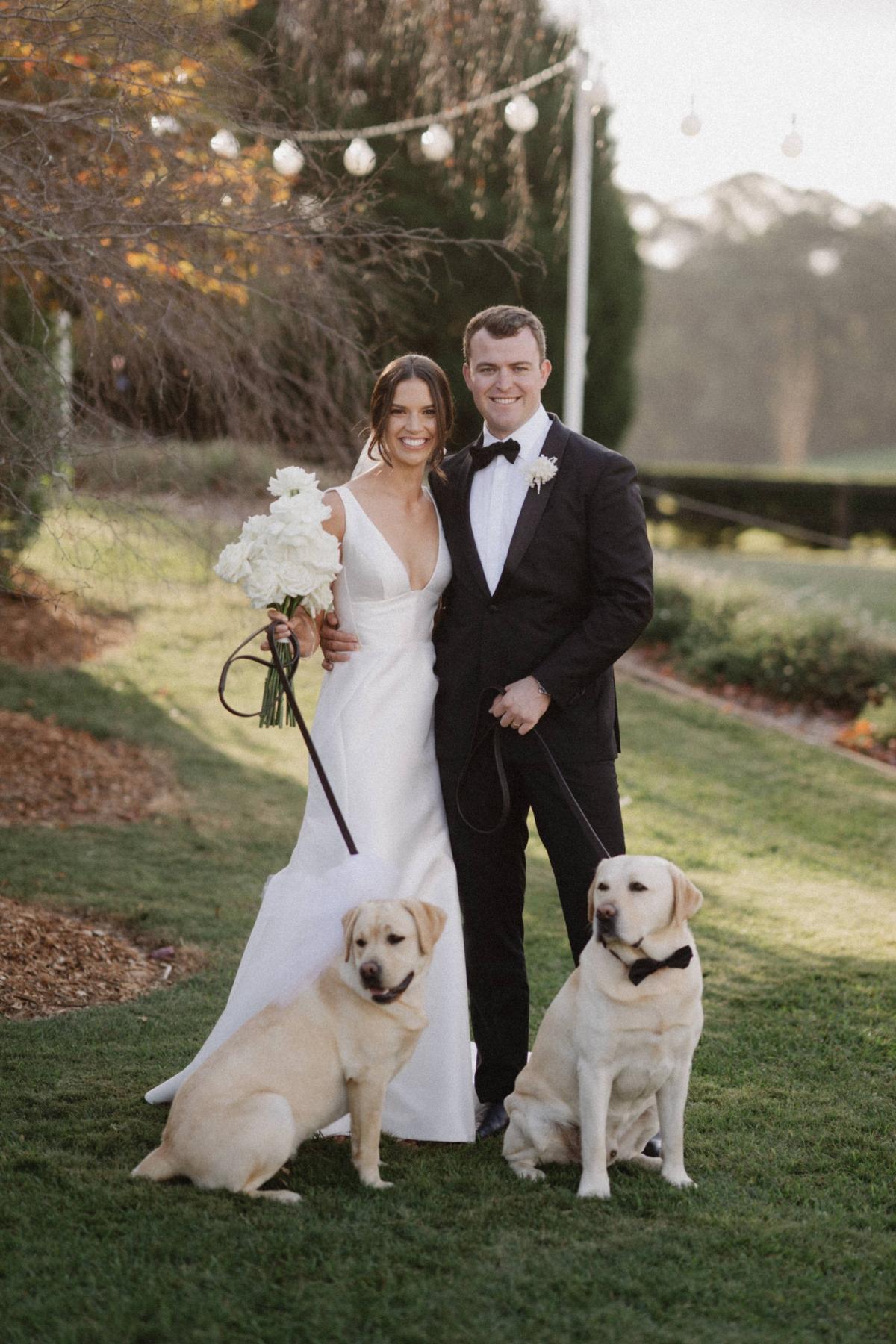 Real bride Charlotte and Myles taking photos with their two blond labs who are wearing black bowties. She wears the elegant Leonie Alexia gown by KWH.