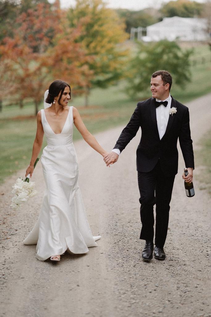 Real bride Charlotte holds hands with Myles while walking down a road together. She wears the modern Leonie Alexia gown by KWH.