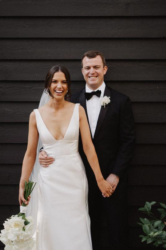 Real bride Charlotte with her new husband Myles standing in front of a black wall. She wears the effortless satin Leonie Alexia gown by KWH