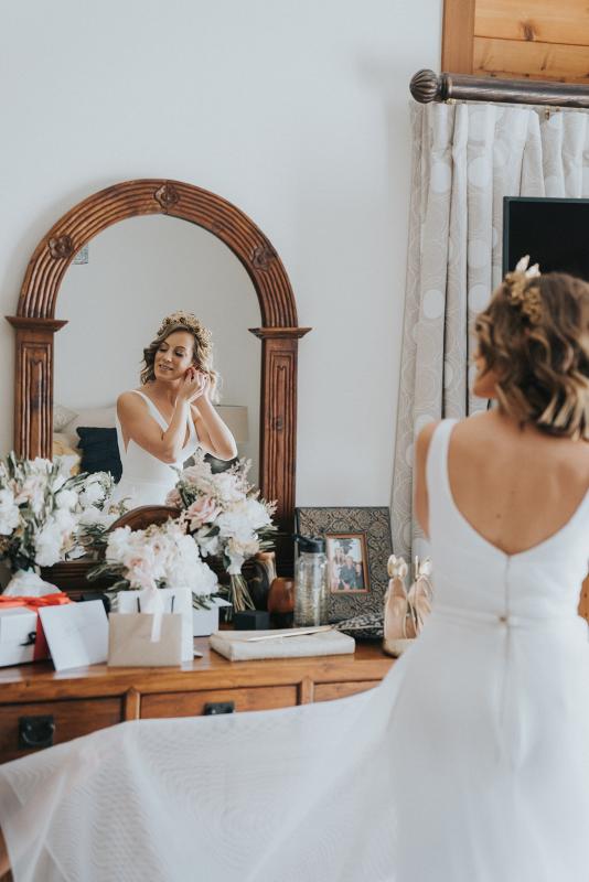 KWH real bride Talia puts in her earrings as she looks in the mirror. She wears the stunning Aisha wedding dress with u-shaped back and satin belt.
