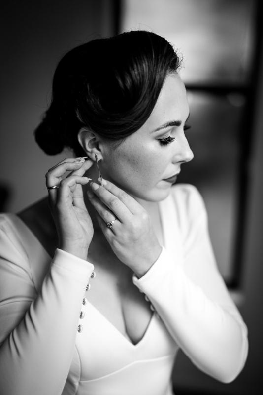 B&W photo of KWH real bride Jaci putting in her earrings. She wears the vintage inspired Aubrey wedding dress with low back and long train.