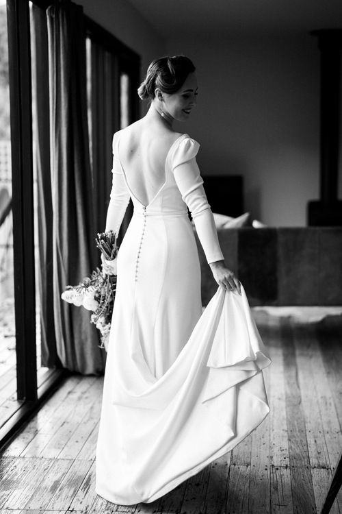 B&W image of KWH real bride Jaci holding the train of her Aubrey wedding dress. It also features a low back and long sleeves.