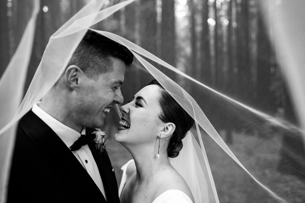 B&W upclose image of KWH real bride Jaci smiling at her husband under her tulle veil. She wears the simple Aubrey wedding dress with v-neck and long sleeves