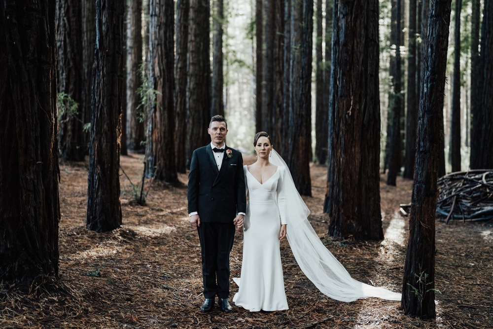 KWH real bride Jaci and Rick stand holding hands in the forest. She wears the ivory crepe Aubrey wedding dress with tulle veil.