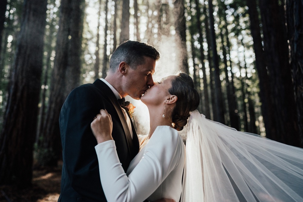 KWH real bride Jaci kisses Rick in the woods. She wears the long sleeved Aubrey wedding dress made from a stretch crepe.