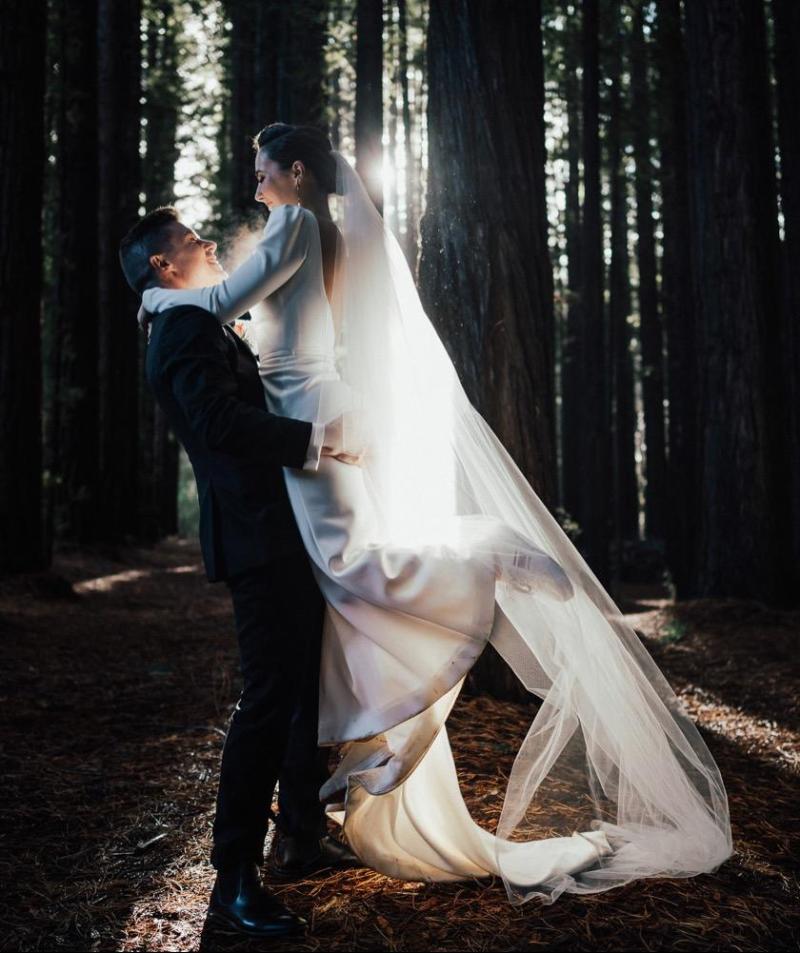 KWH real bride Jaci being pick up and spunin the woods with her new husband. She wears the comfortable Aubrey wedding dress made from crepe with a fitted silhouette.