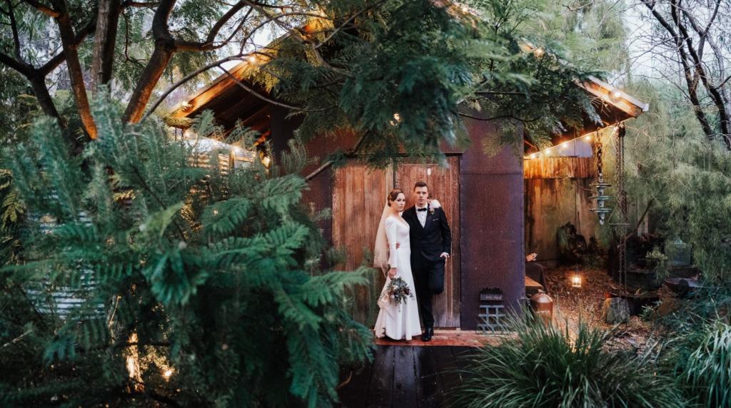 KWH real bride Jaci and Lex standing by a cabin in the woods as she wears the classic Aubrey wedding dress with long sleeves and low back.