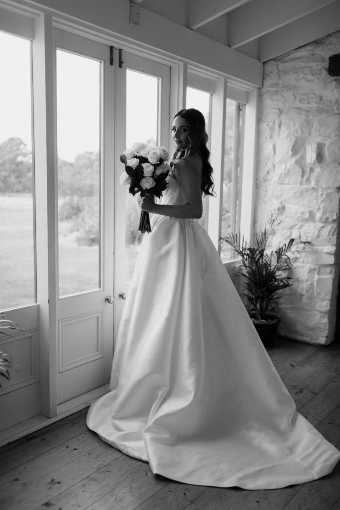 KWH real bride Elke stands in front of the window with her brdial bouquet and ivory Blake Camille wedding dress which features a long train and strapless bodice