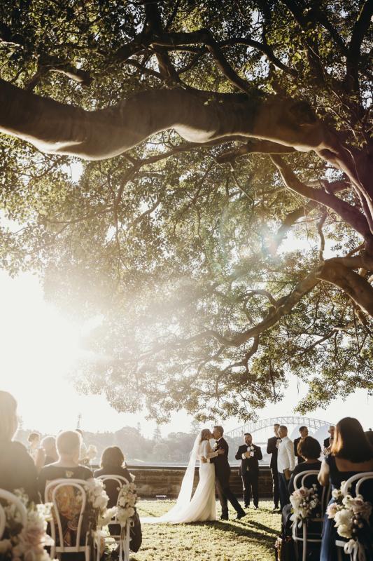 KWH real bride Emily and Mark kissing under a massive tree as the sunsets. She wears the sophisticated Celine wedding dress with sheer beaded sleeves