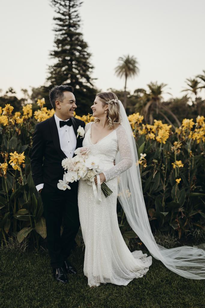 KWH real bride Emily with Mark in front of yellow flowers. She wears the elegant Celine wedding dress with beaded sleeves.