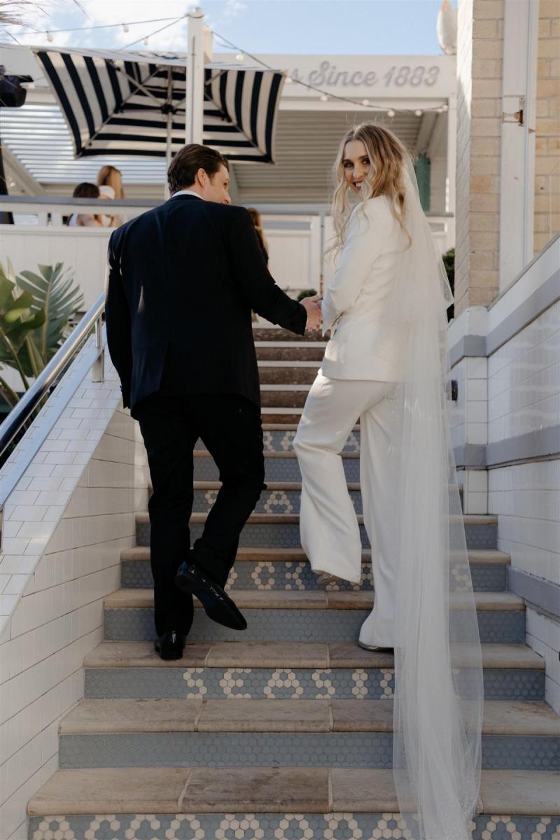 KWH real bride Demi and James walk up greecian looking stairs. She wears the timeless Charlie Danielle bridal suit with her long Astra veil with small sequins.