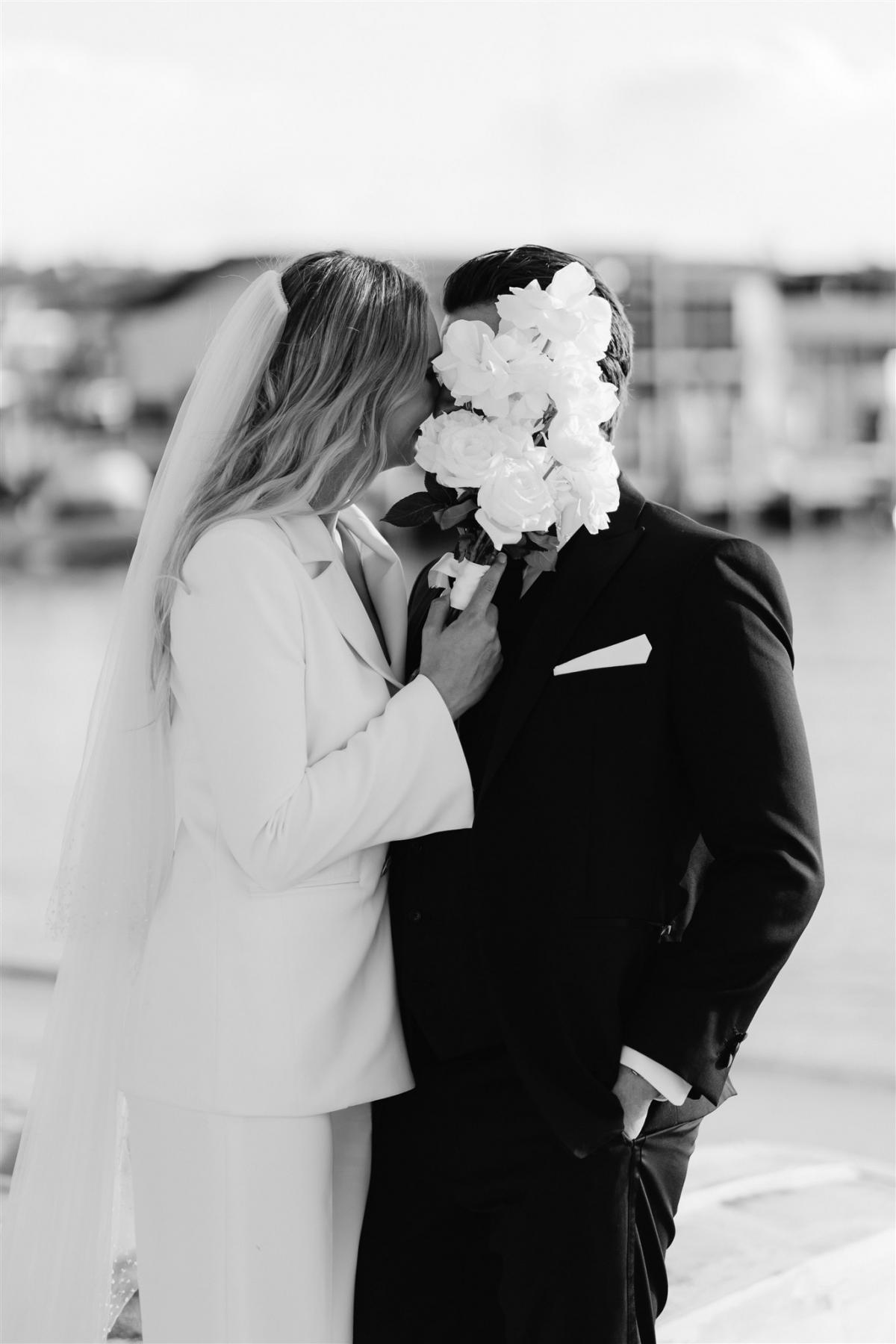 B&W photo of KWH real bride Demi and James kiss behind her white rose bouquet. She wears the stunning Charlie Danielle bridal suit with classic features.