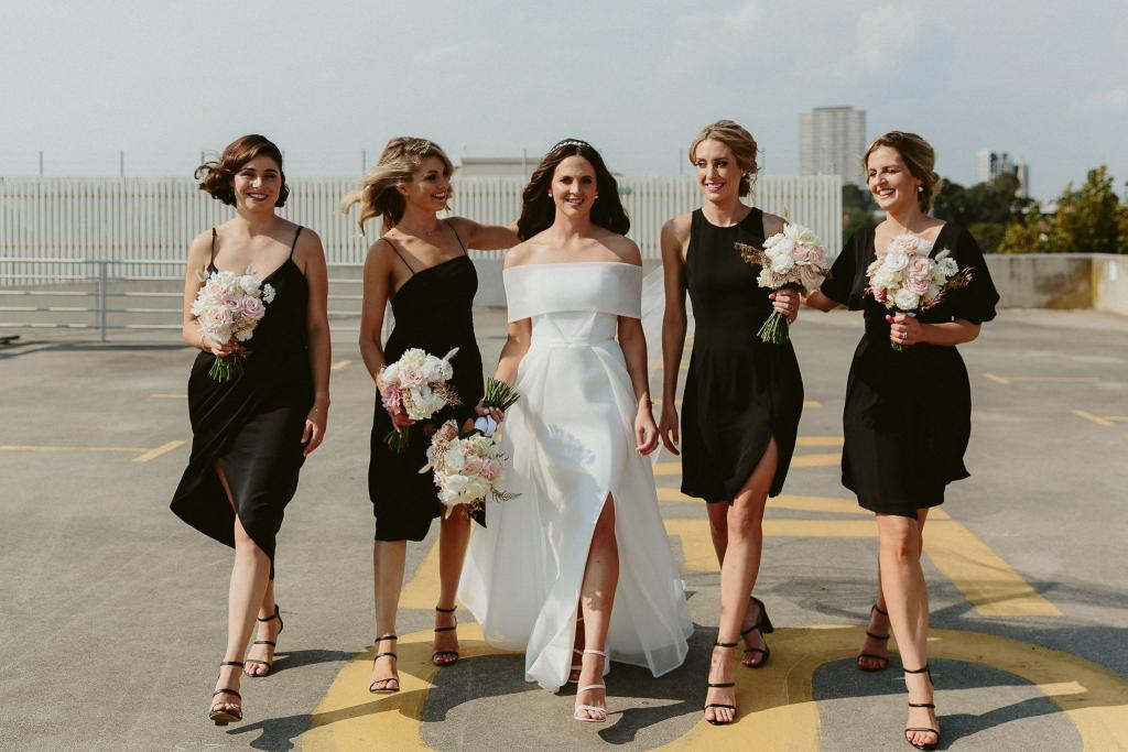 KWH real bride Libby walks in a parking lot with her bridesmaids who wear their black Shona Joy dresses. She wears the ivory Esther wedding dress featuring a front leg split and center pleating