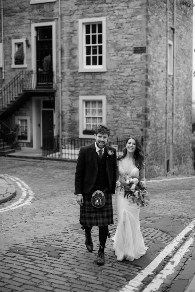 B&W photo of Mel and Robbie walking down an old Edinburgh street hand in hand. She wears the minimalist Jemma wedding dress with fitted skirt and cap sleeves
