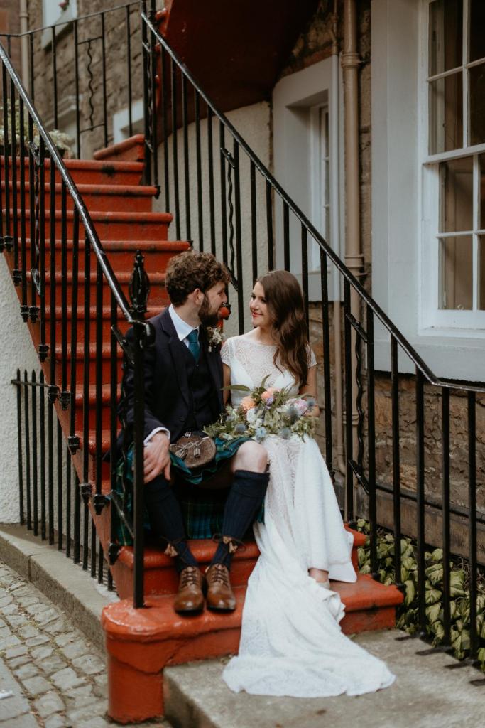 KWH real bride Mel and Robbie sit on an Edinburgh staircase. She wears the classic Jemma wedding dress with illusion neckline and open back.
