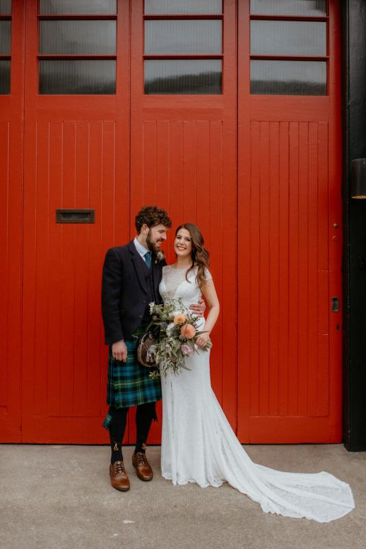 KWH real bride Mel stands in front of a bright red door with Robbie who wears a kilt. She dons the minimalist Jemma wedding dress with all over moder lace.