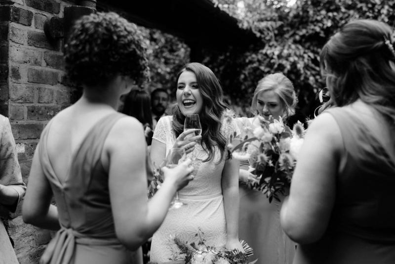 KWH real bride Mel laughing with her bridesmaids as they sip champagne. She wears the fitted lace Jemma wedding dress.