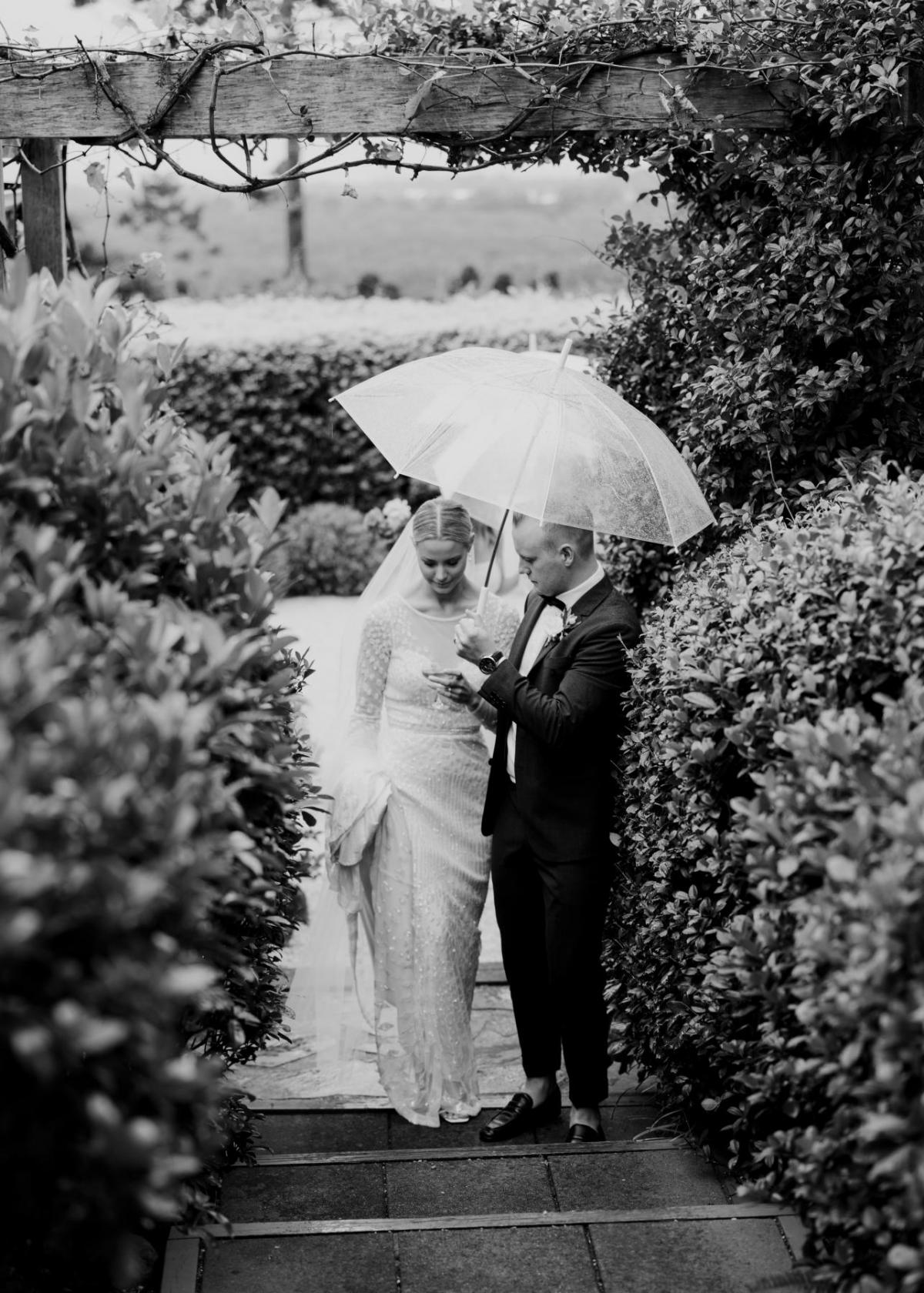 B&W image of Taylor and Kalen under an umbrella as she sheilds her modern Lexie wedding dress with long sleeves