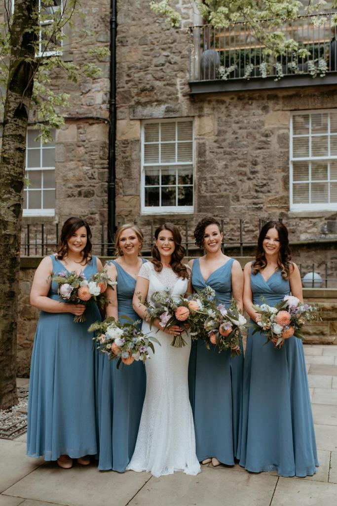 KWH real bride Mel stands with her bridesmaids who are wearing gorgeous blue dresses. She wears the fit and flare Jemma wedding dress made from modern lace.