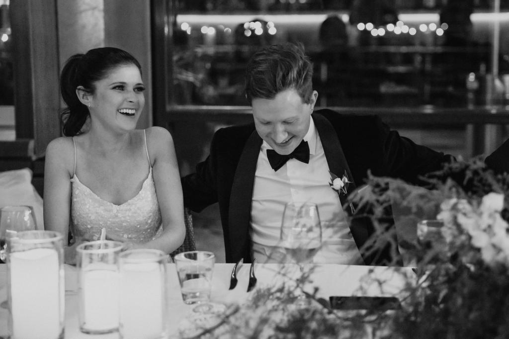 B&W photo of KWH real bride Ash & Chris laughing at their recpetion table. She wears the modern hand beaded Anya wedding dress with shoestring straps.
