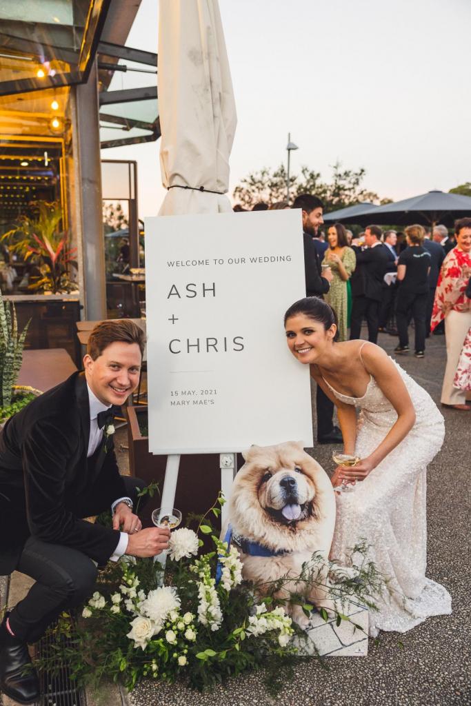 KWH real bride Ash posing with her new husband and Chow Chow by their wedding signage. She wears the sleek Anya wedding dress with form fitting figure.