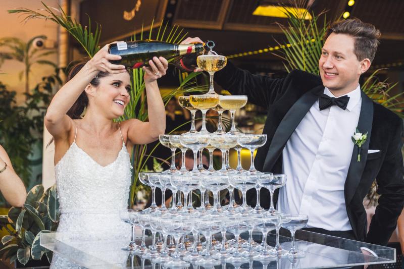 KWH real bride Ash pouring champagne down a glass tower. She wears the sequin Anya wedding dress with short train.