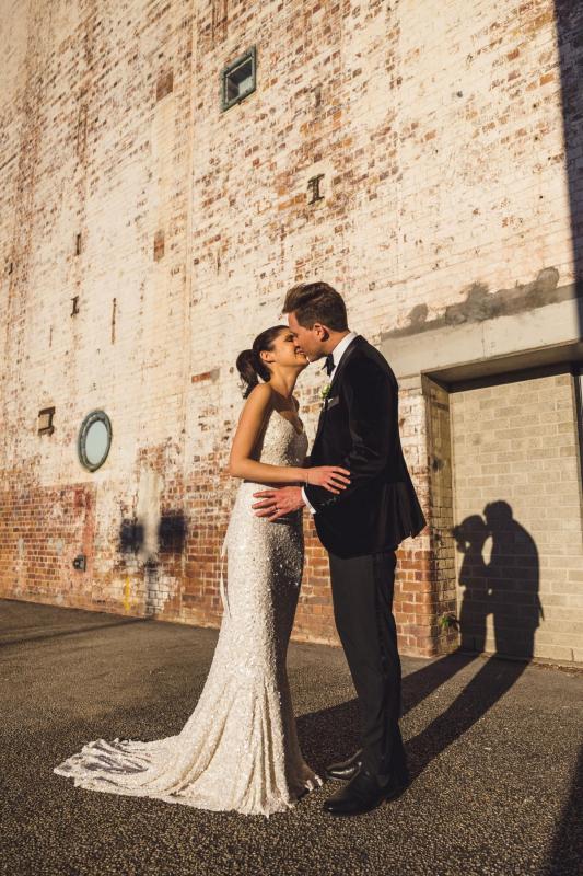 KWH real bride Ash kissing her man in front of a distressed brick building. She wears the sparkley Anya wedding dress with all over sequins.