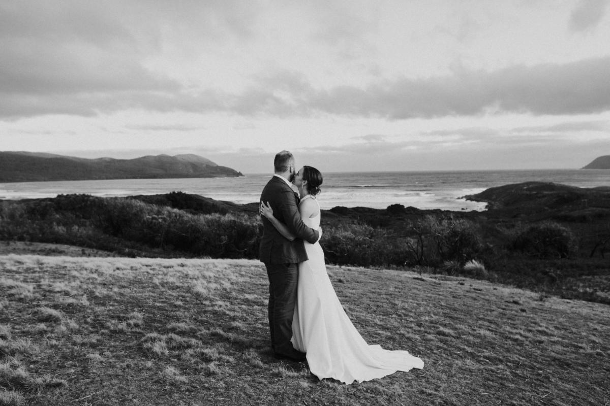 BW image of KWH real bride Ashleigh kissing her new husband against a oceanic backdrop. She wears the minimalist Bridget dress with cutouts on the side.
