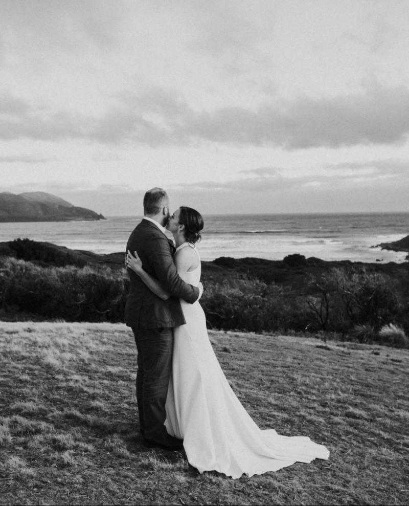 BW image of KWH real bride Ashleigh kissing her new husband against a oceanic backdrop. She wears the minimalist Bridget dress with cutouts on the side.