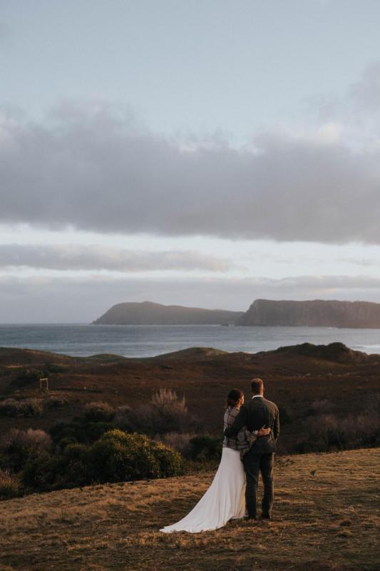 KWH real bride Ashleigh and Simon look over the horizon as the sun sets. She wears the minimalist Bridget wedding dress with racer-back.