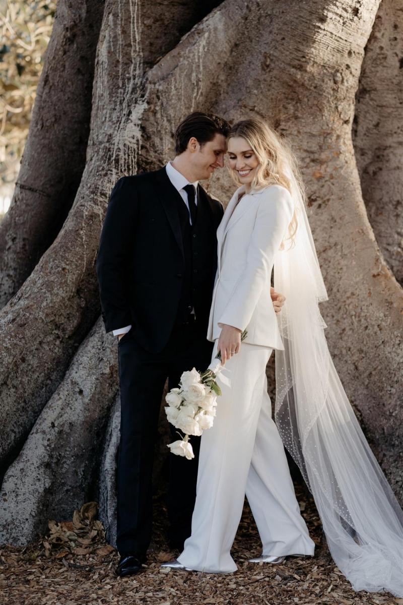 KWH real bride Demi and James stand by a large tree. She wears the classic Charlie Danielle bridal suit with double breasted jacket.